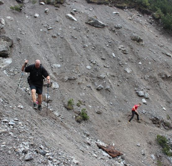 Drei Wanderer steigen einen felsigen Bergpfad hinauf, einer in der Führung mit Trekkingstöcken.
