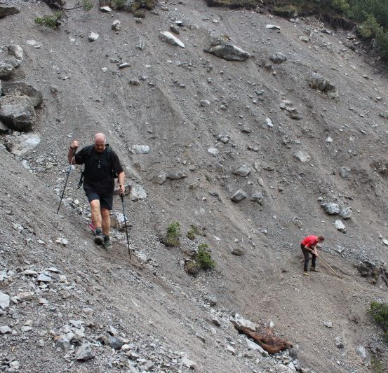 Drei Wanderer steigen einen felsigen Berghang hinauf. Der führende Mann benutzt Trekkingstöcke. Zwei andere sind dahinter, einer hält einen Stock.