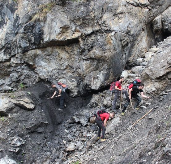 Fünf Personen wandern einen felsigen Berghang hinauf, tragen Rucksäcke und halten Schaufeln. Sie sind in roten Shirts und schwarzen Hosen gekleidet.