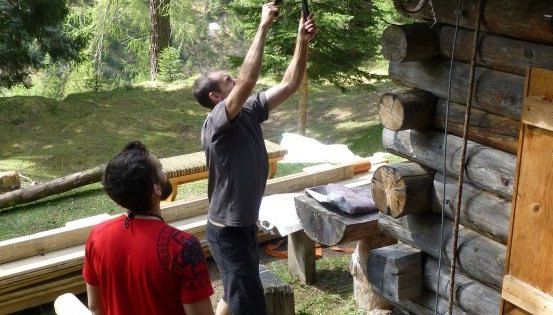 Zwei Männer arbeiten an einer Blockhütte, einer hält ein Werkzeug und der andere beobachtet.