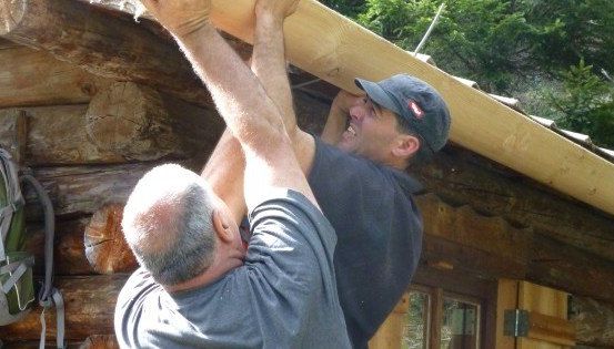 Zwei Männer arbeiten auf dem Dach einer Holzhütte, einer hält einen Holzbalken, während der andere zusieht.