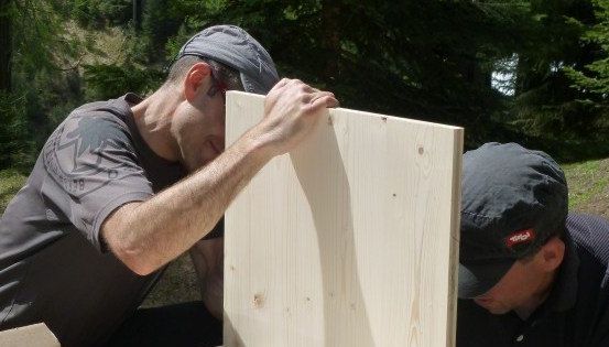 Zwei Männer arbeiten an einer Holzplatte, einer hält sie hoch, während der andere sie genau betrachtet. Sie sind im Freien, umgeben von Bäumen.