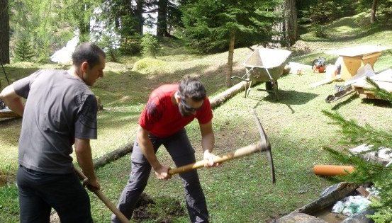 Zwei Männer arbeiten in einem Grasbereich, einer hält eine Spitzhacke und der andere eine Schaufel. Sie sind von verschiedenen Werkzeugen und Materialien umgeben.
