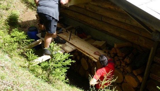 Zwei Männer arbeiten an einer Holzstruktur, einer steht auf einer Planke und der andere ist in einem Lagerschuppen voller Brennholz.