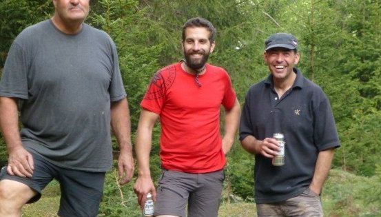 Drei Männer stehen in einem Wald, einer hält eine Bierdose, ein anderer lächelt und der dritte hat einen Bart.