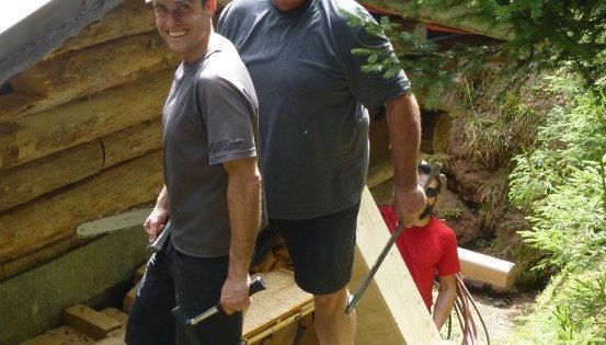 Zwei Männer stehen in einem Wald, einer hält eine Säge und der andere einen Hammer, lächelnd. Hinter ihnen arbeitet eine dritte Person in einem roten Shirt an einer Holzstruktur.