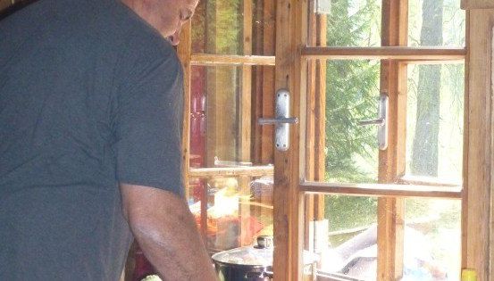 Ein Mann in einem blauen T-Shirt kocht in einer Küche mit einem Fenster, das einen Wald draußen zeigt.