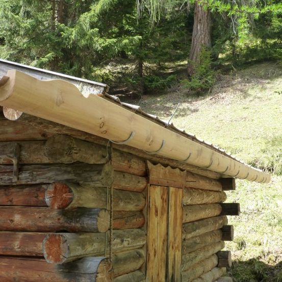 Eine Holzblockhütte mit einem Metalldach und einem Holzdachrinnen. Die Hütte ist von einem Grasbereich mit Bäumen umgeben.