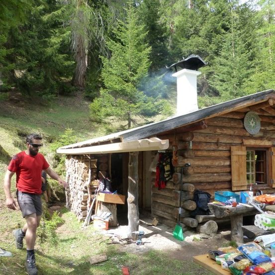 Ein Mann in einem roten T-Shirt und Sonnenbrille geht an einer kleinen Holzhütte mit Solarpanel und Rauch aus dem Schornstein vorbei.