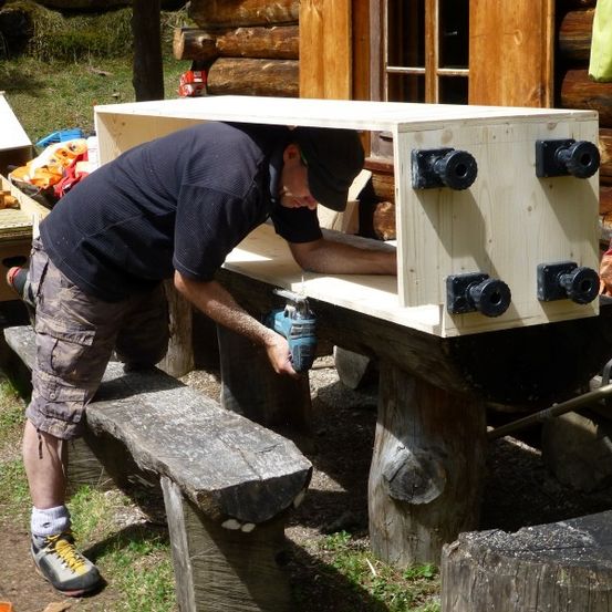 Ein Mann arbeitet an einer Holzkiste auf einer Bank vor einer Blockhütte. Er benutzt eine Bohrmaschine und hat Werkzeuge und Materialien um sich herum.