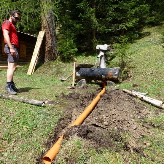 Ein Mann in einem roten Shirt und schwarzen Shorts steht neben einer großen orangefarbenen Rohrleitung, die im Boden vergraben ist. Hinter ihm befindet sich eine Holzstruktur mit einer Wasserpumpe.