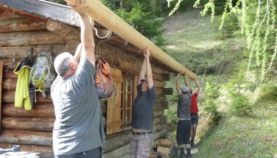 Vier Männer arbeiten gemeinsam daran, einen großen Holzbalken auf das Dach einer Blockhütte zu heben. Die Hütte ist von Grün umgeben.