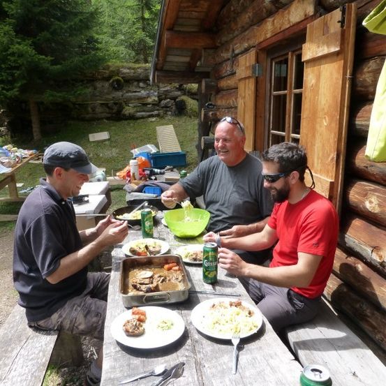 Drei Männer sitzen an einem Tisch vor einer Blockhütte und essen und trinken Dosengetränke.