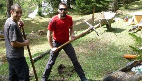 Zwei Männer stehen in einem Grasbereich, einer hält einen langen Stock und der andere eine große Axt. Hinter ihnen steht eine Schubkarre, eine Holzkonstruktion und ein Holzstamm.