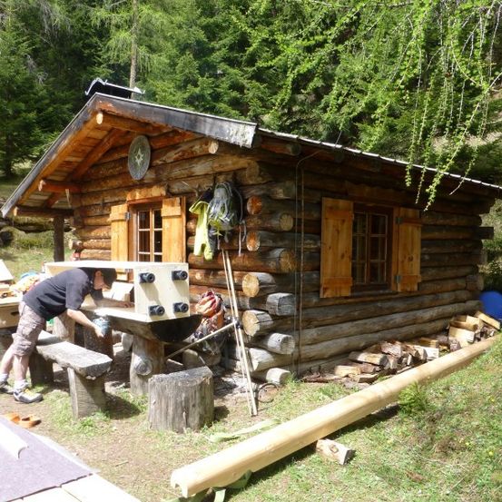 Ein Mann arbeitet an einer Holzbank vor einer Blockhütte, umgeben von Holz und Grün.