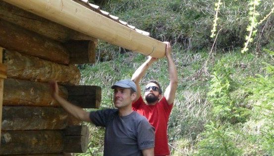 Zwei Männer arbeiten an einer Holzstruktur, einer hält einen Holzbalken, während der andere ihn von unten stützt. Sie sind im Freien, umgeben von Grün.