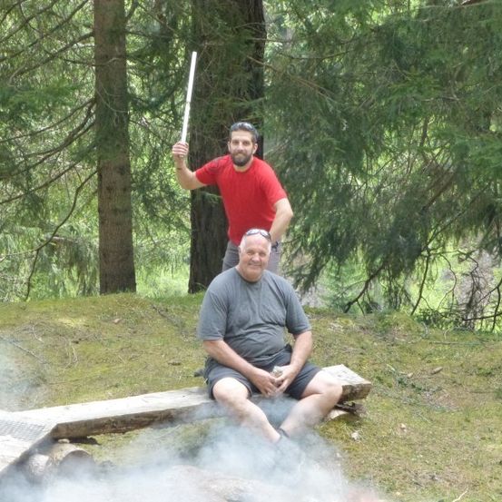Zwei Männer sitzen auf einer Bank im Wald, einer hält einen Stock. Der Mann auf der rechten Seite lächelt.