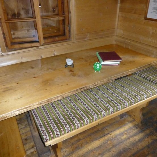 Ein hölzerner Tisch mit Bänken und einem Fenster in einem hölzernen Raum. Es gibt Bücher und einen grünen Gegenstand auf dem Tisch.