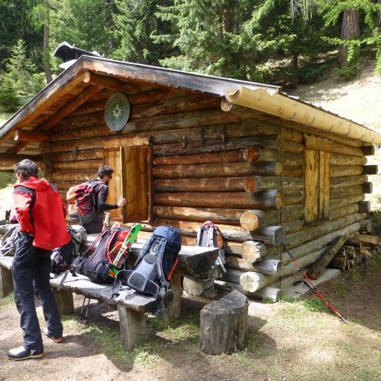 Eine Gruppe von Wanderern steht vor einer kleinen Holzhütte im Wald, mit Rucksäcken und Skiern in der Nähe.