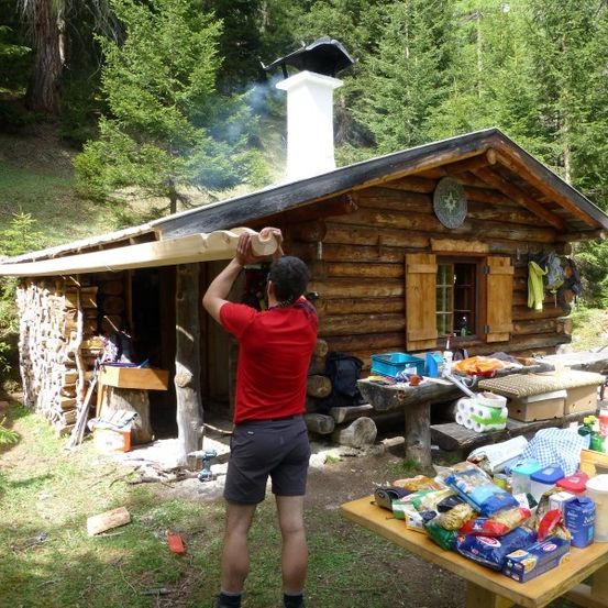 Zwei Männer arbeiten an einer Holzhütte im Wald. Einer hält einen Holzbalken, der andere klettert hinauf. Die Hütte hat einen Schornstein, eine Uhr und eine Tür. Es gibt Bänke, einen Tisch und verschiedene Gegenstände auf dem Boden.
