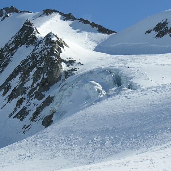 Ein Gebirgszug mit schneebedeckten Gipfeln und einem Gletscher im Vordergrund. Der Himmel ist klar und blau.