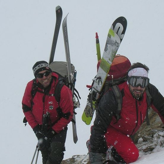 Zwei Männer in roten Jacken und Schutzbrillen wandern einen schneebedeckten Berg hinauf, tragen Skier und Rucksäcke.