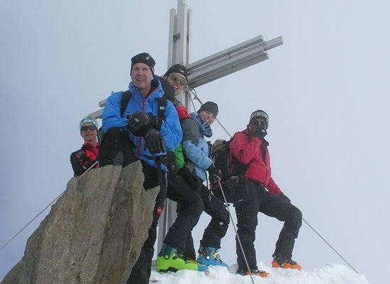 Eine Gruppe von Menschen in Winterkleidung posiert für ein Foto am Gipfel eines verschneiten Berges, mit einem großen Kreuz im Hintergrund.