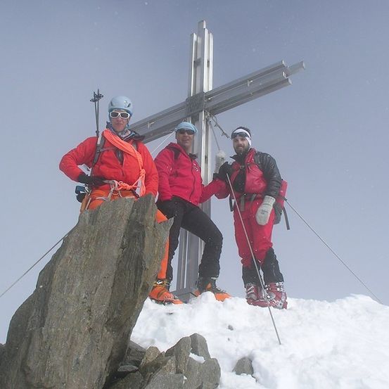 Drei Kletterer in roten Ausrüstungen stehen auf einem verschneiten Berg und posieren mit einem großen Kreuz im Hintergrund.