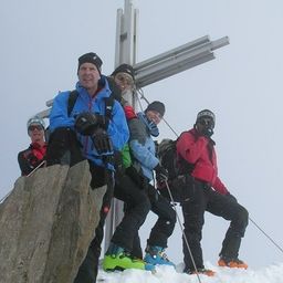 Eine Gruppe von Wanderern posiert für ein Foto auf einem verschneiten Berggipfel mit einem Kreuz im Hintergrund.