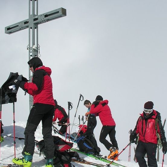 Eine Gruppe von Skifahrern in roten Jacken und schwarzen Hosen bereitet sich auf eine Skitour auf einem verschneiten Berg vor. Sie stehen in der Nähe eines Kreuzes und einige passen ihre Skiausrüstung an.
