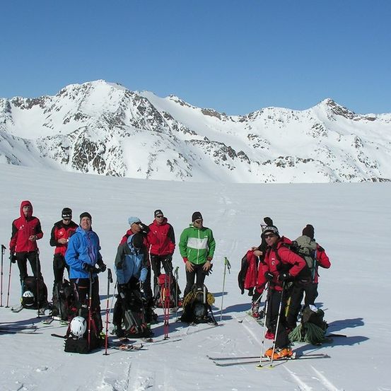 Eine Gruppe von Skifahrern steht in einer verschneiten Landschaft, mit Bergen im Hintergrund und einem klaren blauen Himmel.