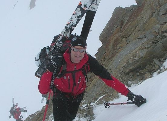 Ein Mann in einem roten Jackett und schwarzem Hut klettert einen verschneiten Berg hinauf, Skier und Stöcke im Gepäck. Er lächelt und scheint den Aufstieg zu genießen.