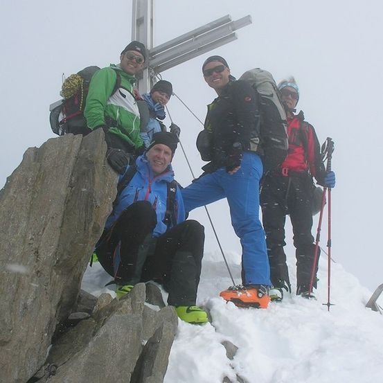 Fünf Personen in Winterausrüstung posieren auf einem verschneiten Berggipfel, einer sitzt auf einem Felsen, während andere hinter ihm stehen.