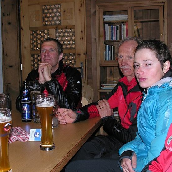 Eine Gruppe von Menschen sitzt um einen Tisch mit Biergläsern, ein Mann trägt eine schwarze Lederjacke und ein anderer eine rote Jacke. Hinter ihnen eine Holztür mit einem Glasfenster und ein Holzschrank mit Büchern.