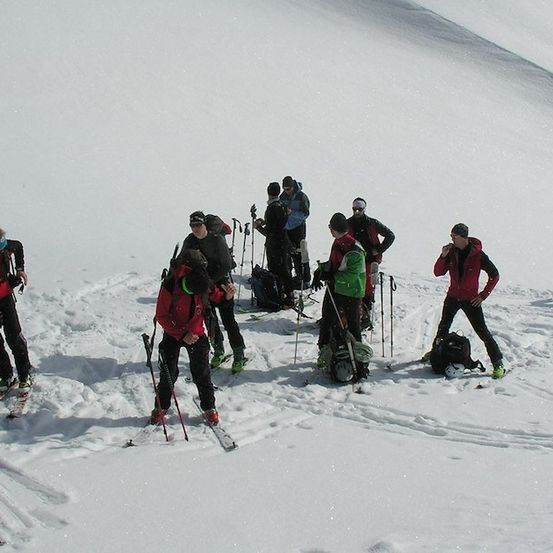 Eine Gruppe von Skifahrern versammelt sich auf einem verschneiten Berg, einige stehen und andere bereiten ihre Ausrüstung vor. Sie sind in Winterkleidung gekleidet und halten Skistöcke. Der Schnee ist weiß und der Himmel klar.