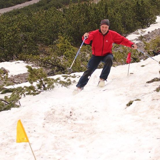 Ein Mann in einem roten Jackett schneidet auf einem verschneiten Hang, hält Skistöcke und lächelt.