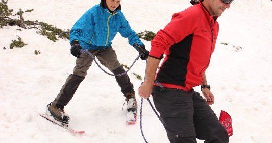 Ein Mann und ein Kind üben Langlauf auf einem verschneiten Berg, verbunden durch ein Seil. Das Kind trägt eine blaue Jacke und der Mann eine rote Jacke.