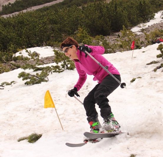 Eine Frau in einer rosa Jacke skifahrert einen schneebedeckten Hang hinunter, ein gelbes Flaggenzeichen ist in der Nähe.