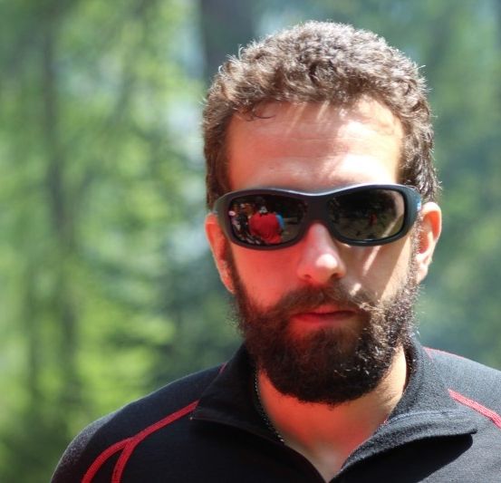 Ein Mann mit Bart, der eine Sonnenbrille und ein schwarzes Shirt mit rotem Rand trägt, steht in einem Wald.