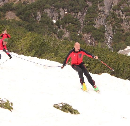 Zwei Männer in roten Skianzügen fahren einen verschneiten Berg hinunter, einer wird vom anderen gezogen.