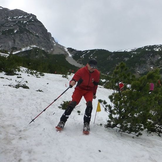 Ein Mann rennt auf einem verschneiten Berg, trägt ein rotes Outfit und hält Skistöcke.