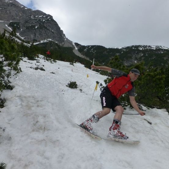 Ein Mann fährt mit Skiern den schneebedeckten Berghang hinunter, trägt eine rote Weste und schwarze Shorts. Er hält einen Skistock in der rechten Hand.