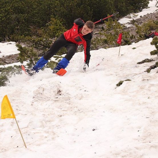 Ein Skifahrer navigiert einen schneebedeckten Hang, trägt ein rotes und schwarzes Outfit und blaue Hosen. Er ist mitten im Sprung, mit umherspritzendem Schnee. Es gibt gelbe Flaggen, die den Weg markieren.