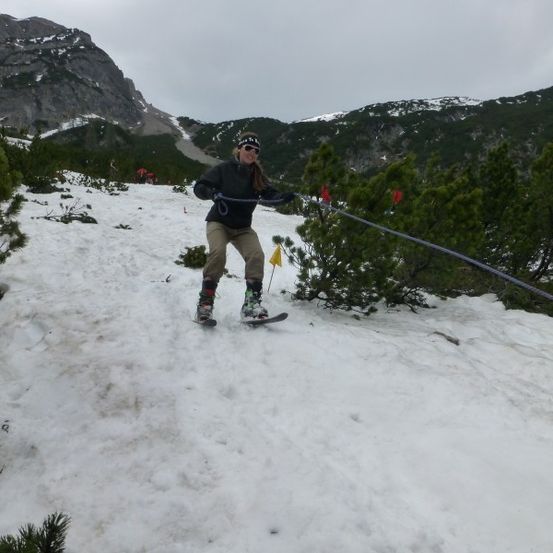 Eine Frau skilanglauft einen schneebedeckten Berg hinauf und hält an einem Seil fest. Sie trägt eine schwarze Jacke, Sonnenbrille und Skischuhe.