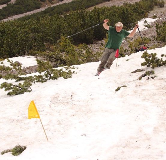 Ein Mann rutscht einen schneebedeckten Hang hinunter, hält Skistöcke und eine gelbe Fahne im Schnee.
