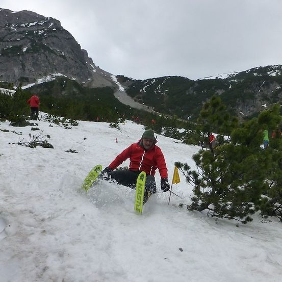 Ein Skifahrer in einem roten Jackett und grünem Hut gleitet einen schneebedeckten Berg hinunter, mit Kiefern und einer gelben Fahne im Hintergrund.