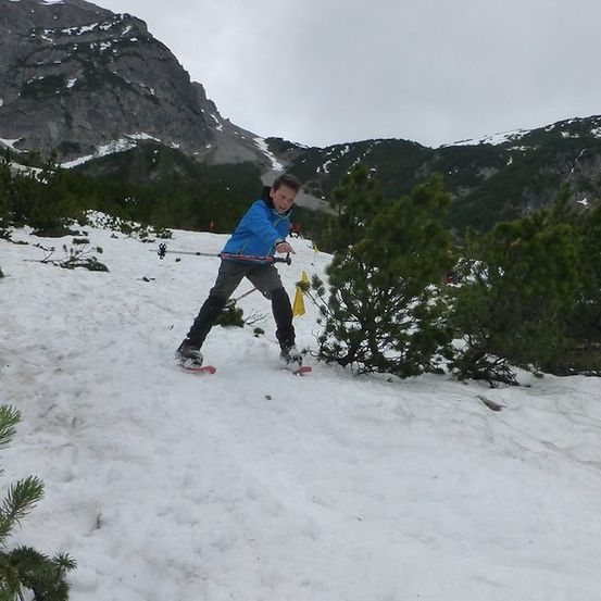 Ein junger Junge skirt einen schneebedeckten Berg hinunter, hält Skistöcke und trägt eine blaue Jacke.