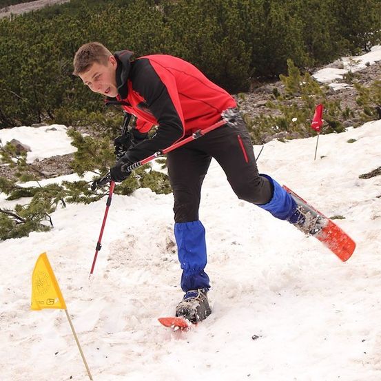 Ein Skifahrer in einem roten Jackett und schwarzen Hosen fährt einen schneebedeckten Hang hinunter und hält Skistöcke.