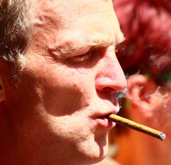 Ein Mann mit grauem Haar raucht einen Zigarren, die Augen geschlossen, und Rauch wirbelt um sein Gesicht.