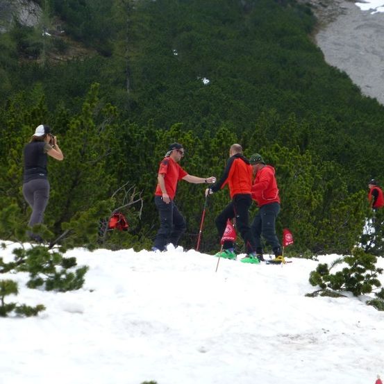 Eine Gruppe von Skifahrern in roten und schwarzen Kleidungsstücken steht auf einem verschneiten Berg, einer hält eine Kamera und ein anderer Skistöcke.